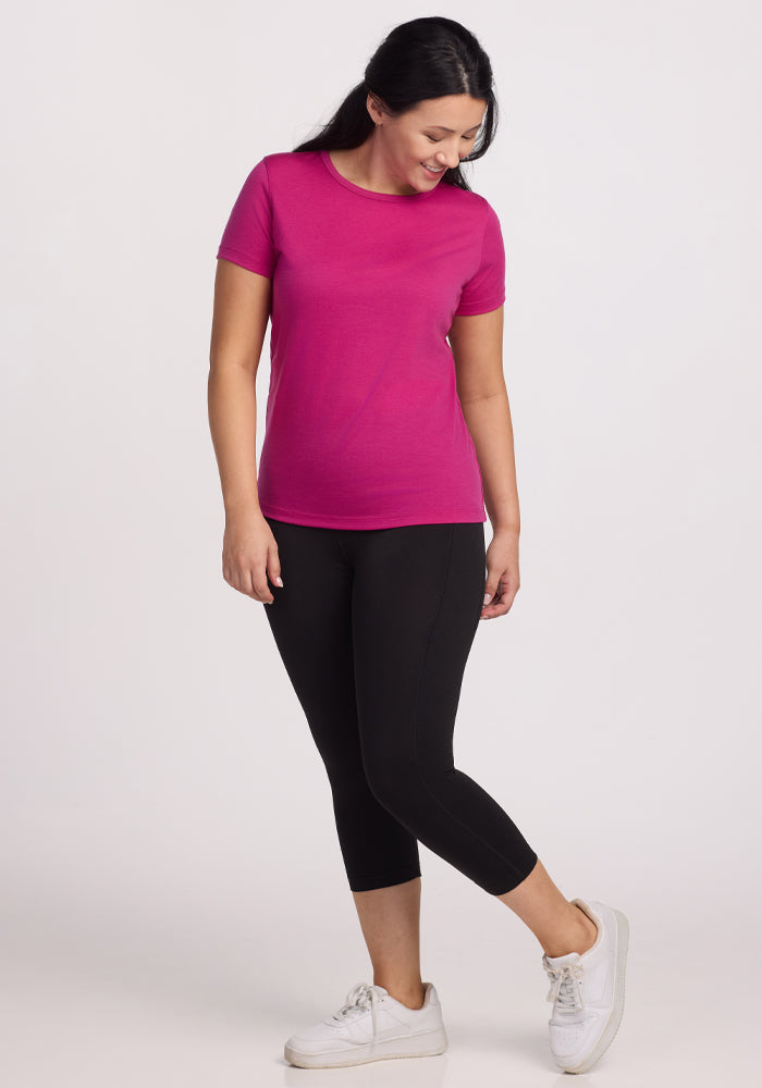 A person wearing the Woolx Liza Crew Neck Tee in Berry Punch, along with black leggings and lightweight white sneakers, stands against a plain background. They're smiling and looking down with one foot slightly raised.