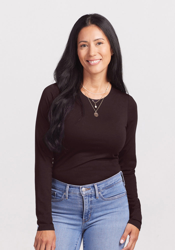 A woman with long black hair, wearing a glamorous Woolx Sophia Bodysuit and light blue jeans, is smiling at the camera. She has a layered necklace with small pendants. The background is plain white. 