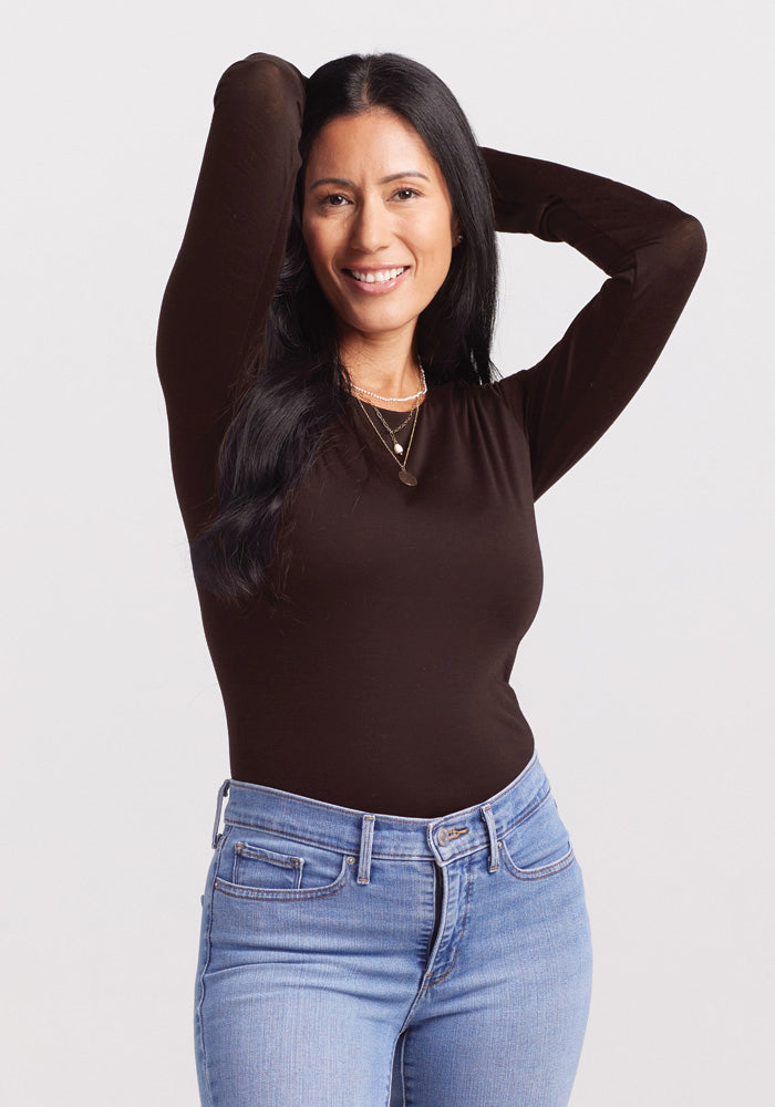 A woman with long dark hair, showcasing a chic look in a Woolx long-sleeved black Sophia Bodysuit paired with light blue jeans, stands with her hands raised and resting on her head. She is smiling and facing the camera against a plain light gray background.