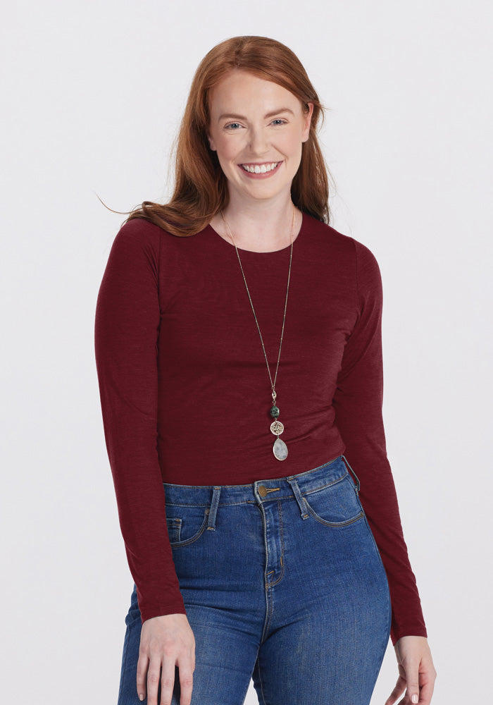 A smiling woman with long auburn hair is wearing a glamorous fit maroon long-sleeve Sophia Bodysuit from Woolx and blue jeans. She has a long necklace with three circular pendants. The background is plain white. 