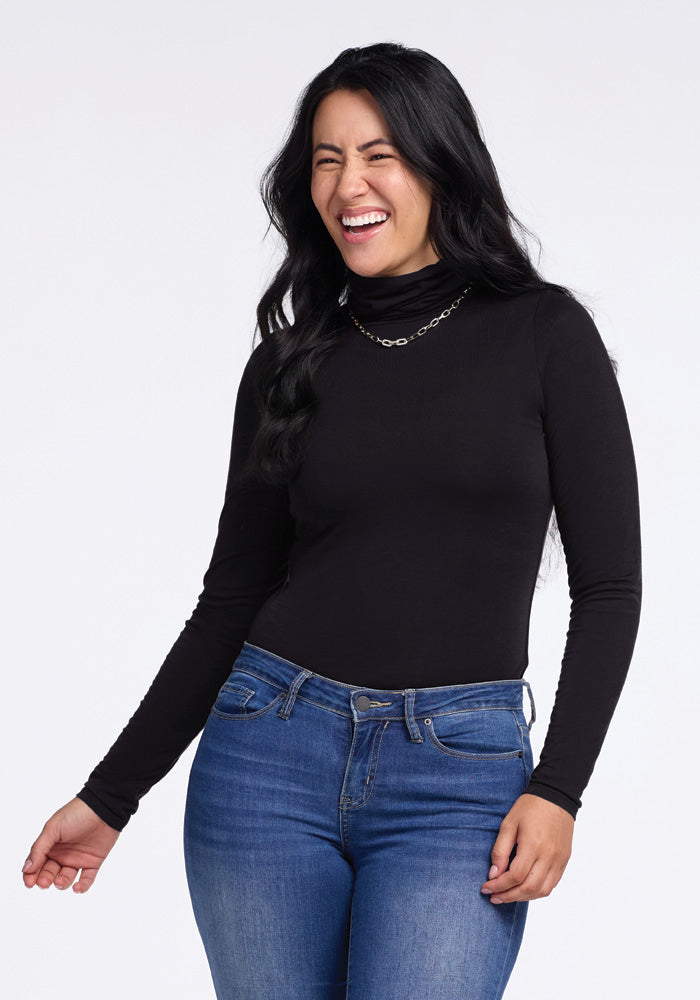 A woman with long dark hair is wearing a Woolx Kennedy Bodysuit in black and blue jeans. She is looking slightly to the side and laughing with her eyes closed. She’s accessorizing with a long silver necklace, and her right arm is bent slightly while her left arm is down by her side.