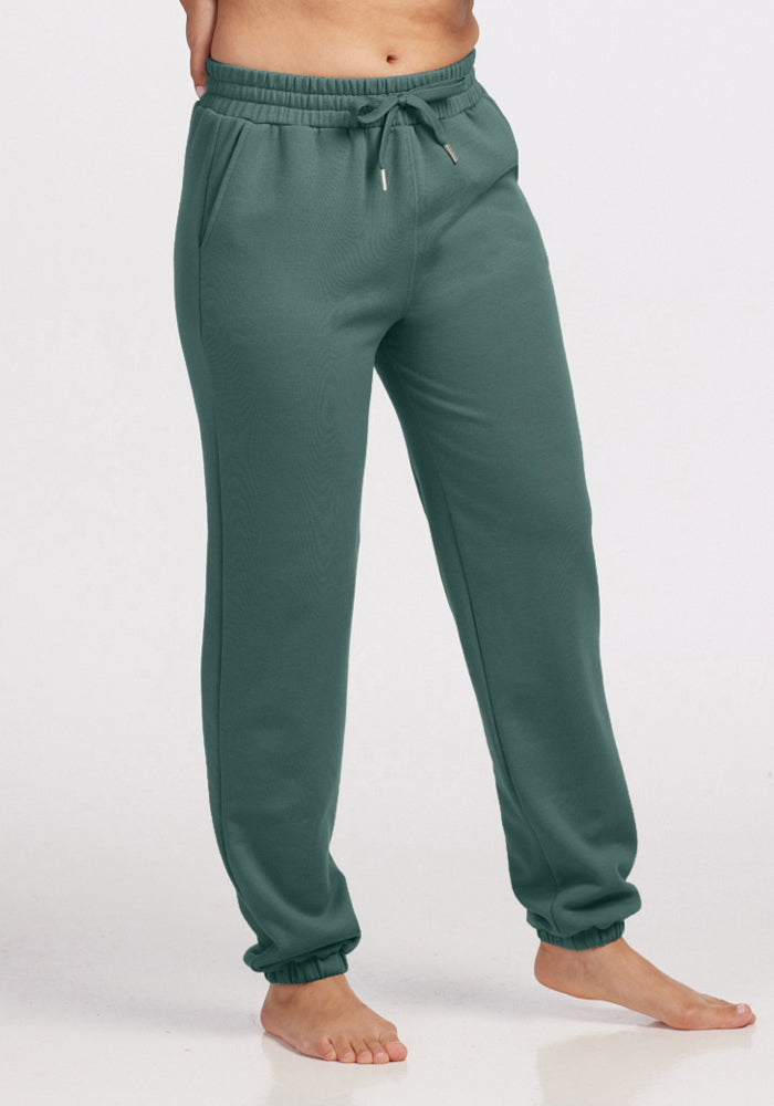 Someone stands barefoot on a light background wearing Woolx's Parker Sweatpants in Duck Green. These temperature-regulating joggers are designed with an elastic waistband and drawstring, featuring elastic cuffs at the ankles, while their upper body remains out of view. 