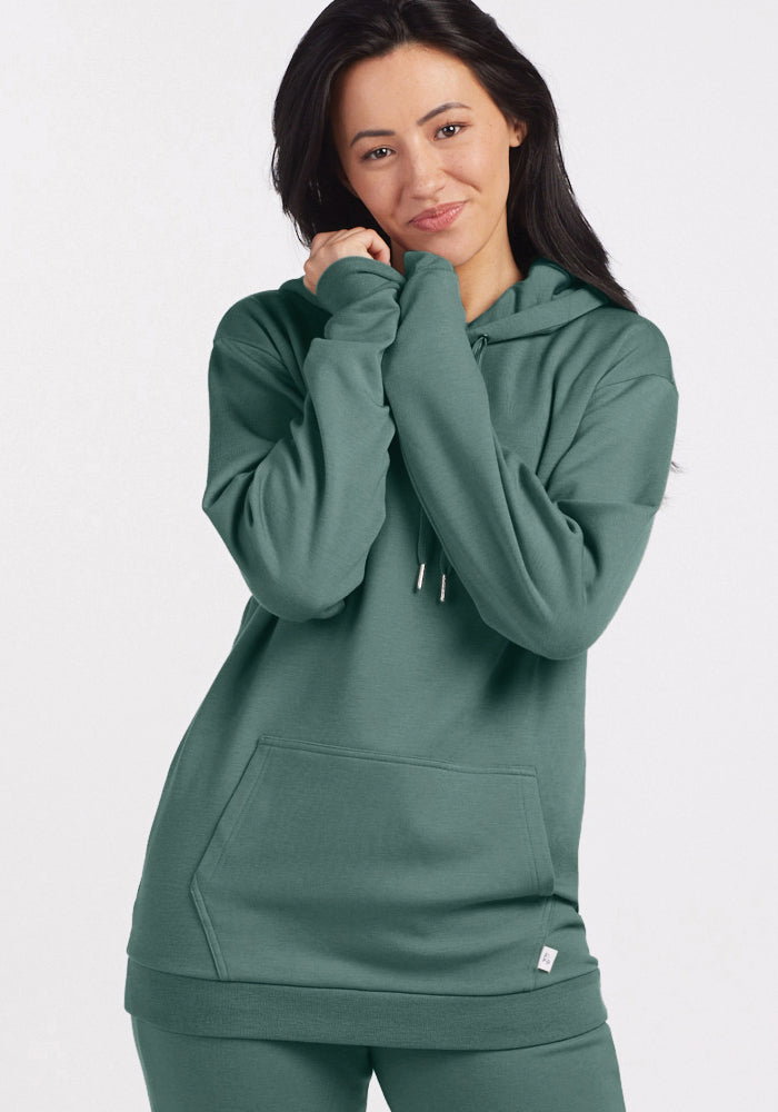 A person with long dark hair wears the Avery Hoodie in Duck Green from Woolx, crafted from soft Merino wool, along with matching pants. They stand against a plain white background, smiling while gently touching their face and neck.