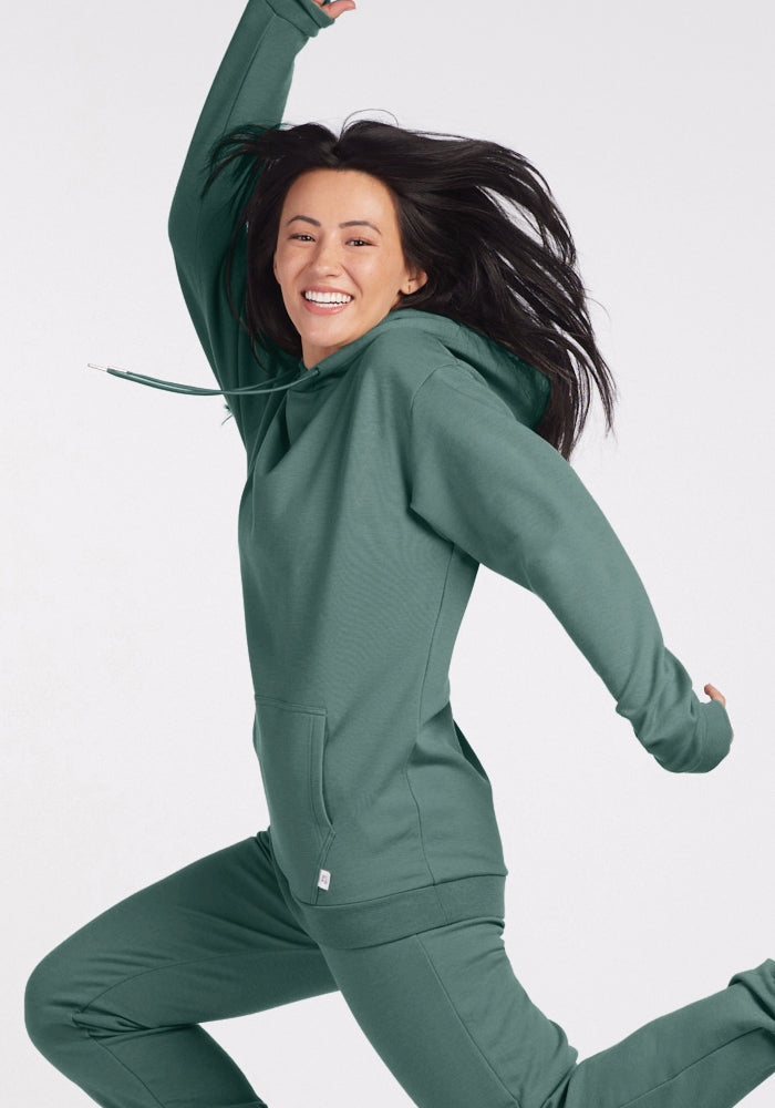 A person wearing the Avery Hoodie in Duck Green from Woolx is joyfully jumping in the air against a plain background, with a big smile and their hair flowing.