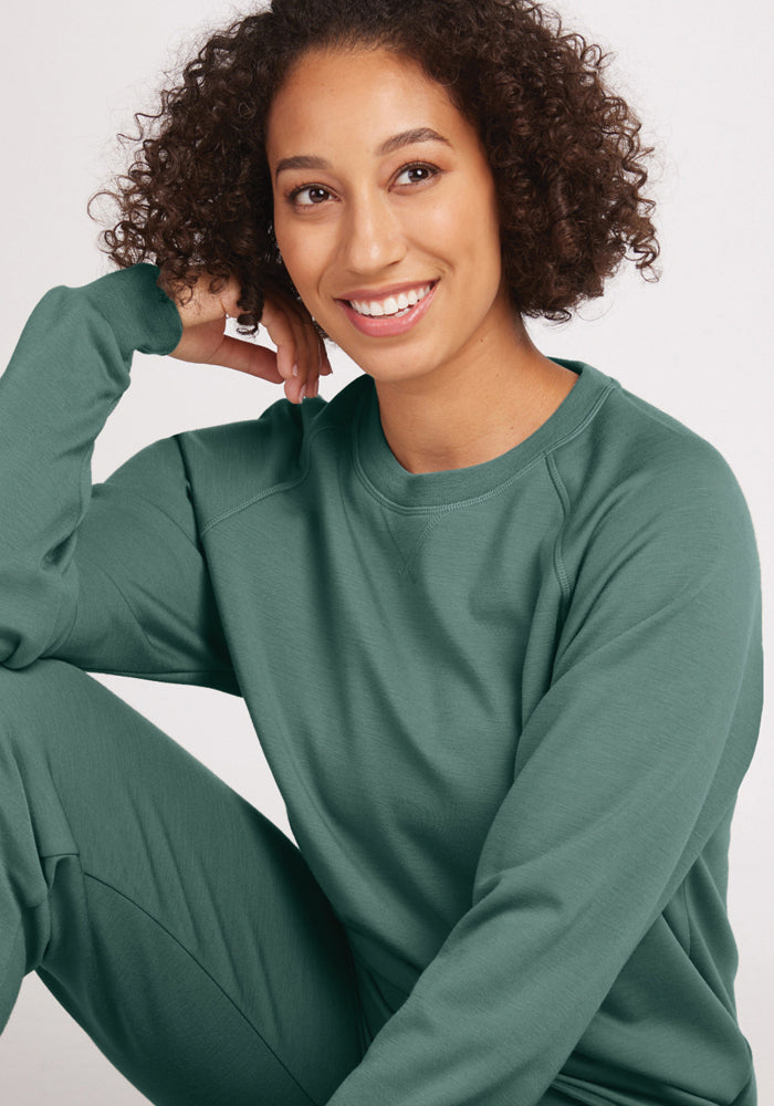 A person with curly hair smiles while wearing the Bailey Crewneck Sweatshirt in Duck Green from Woolx. They are sitting casually with one elbow resting on their knee against a plain, light background.