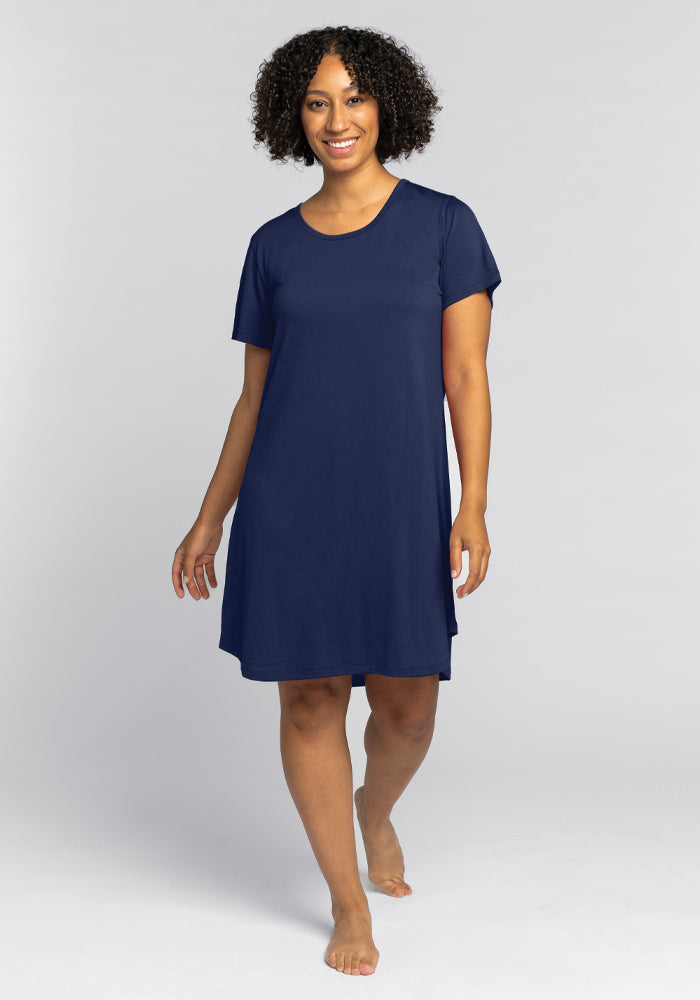 A person with curly hair is wearing a navy blue short-sleeved Desi Nightgown - Starry Night from Woolx and is walking barefoot. The background is a plain light gray.