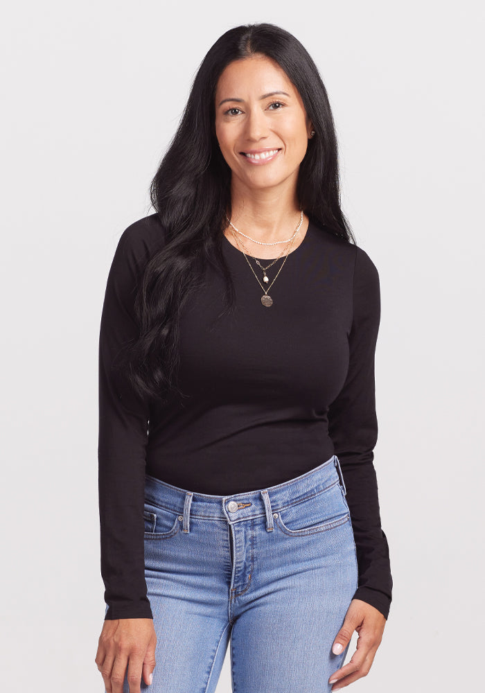 A person with long black hair and a relaxed demeanor wears a Woolx Sophia Bodysuit in black and light blue jeans, smiling against a plain white background. They've layered necklaces for style. 