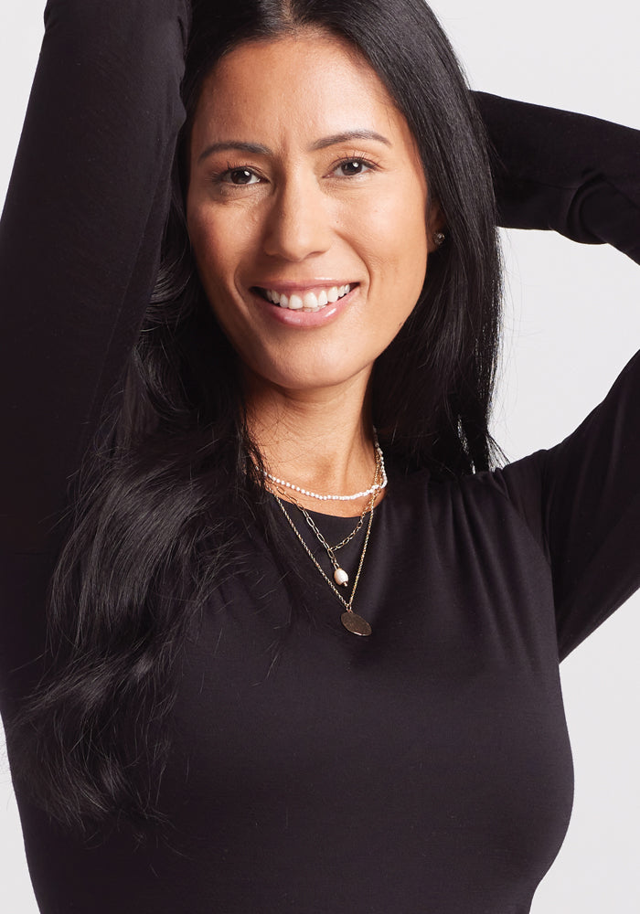 A long-haired woman smiles at the camera, wearing a Woolx Sophia Bodysuit in black and layered necklaces against a plain, light-hued background.