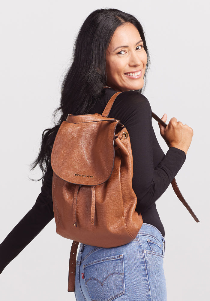 A person with long dark hair smiles while wearing a black Sophia Bodysuit by Woolx and carrying a brown leather backpack. They are turned slightly to the side, showcasing the backpack against a plain white background.