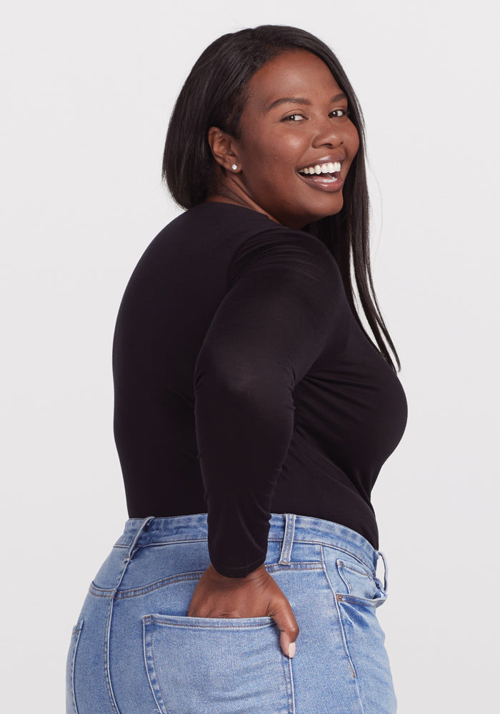 A person with long hair smiles, wearing a black Woolx Sophia Bodysuit and light blue jeans. They stand against a plain white background, slightly turned to the side with one hand in their pocket.