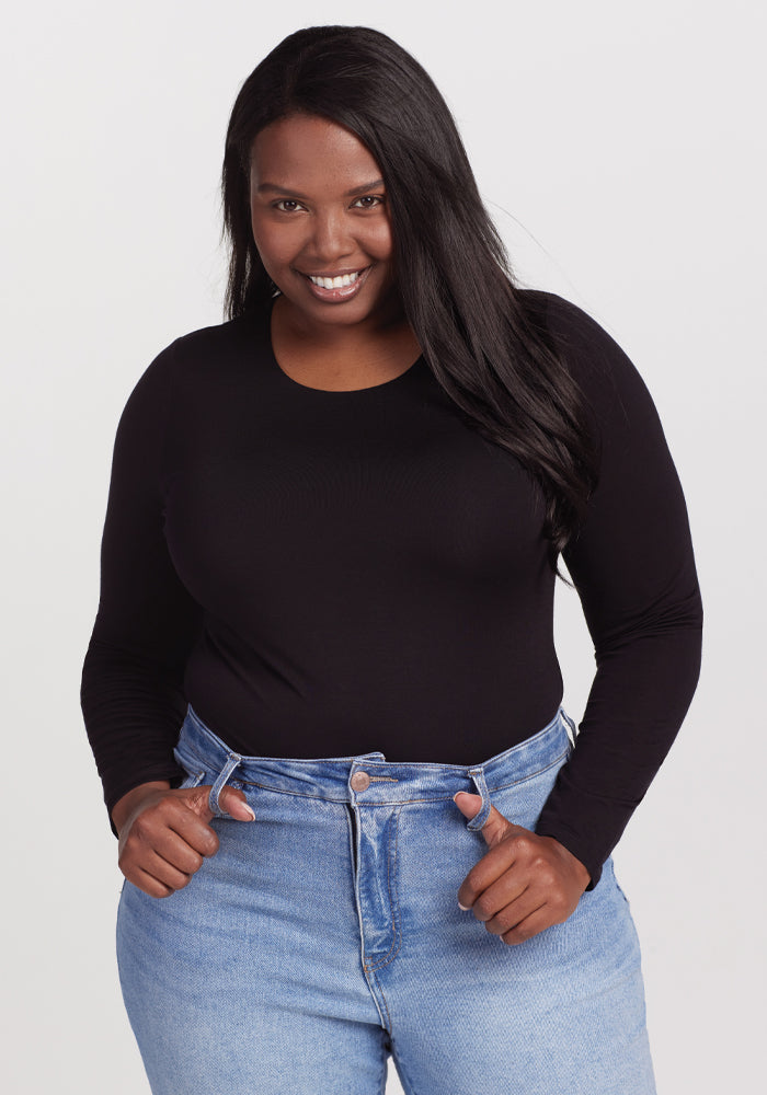 A person smiling, dressed in a Woolx Sophia Bodysuit - Black and high-waisted blue jeans. Their long, dark hair flows naturally as they stand against a simple white background. 