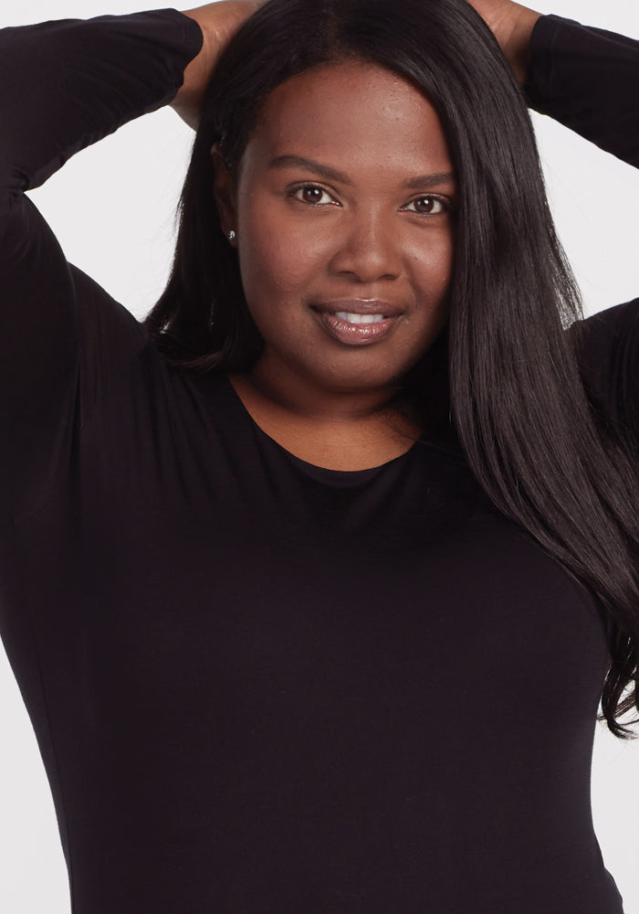 A person with long dark hair smiles gently, posing with their hands on their head. They wear a black Sophia Bodysuit by Woolx, admired for its moisture-wicking properties, against a plain white background.