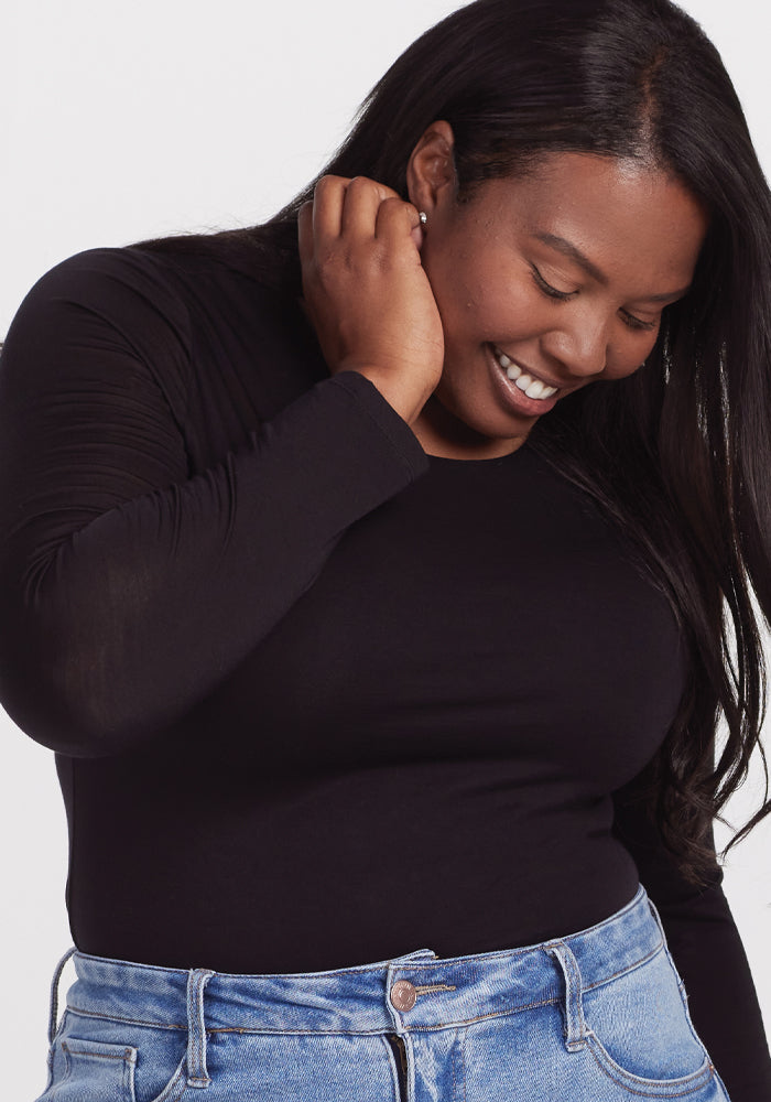 A person with long dark hair smiles while looking down, touching their ear. They are wearing a black Sophia Bodysuit by Woolx made of moisture-wicking Merino wool and blue jeans. The background is light-colored.