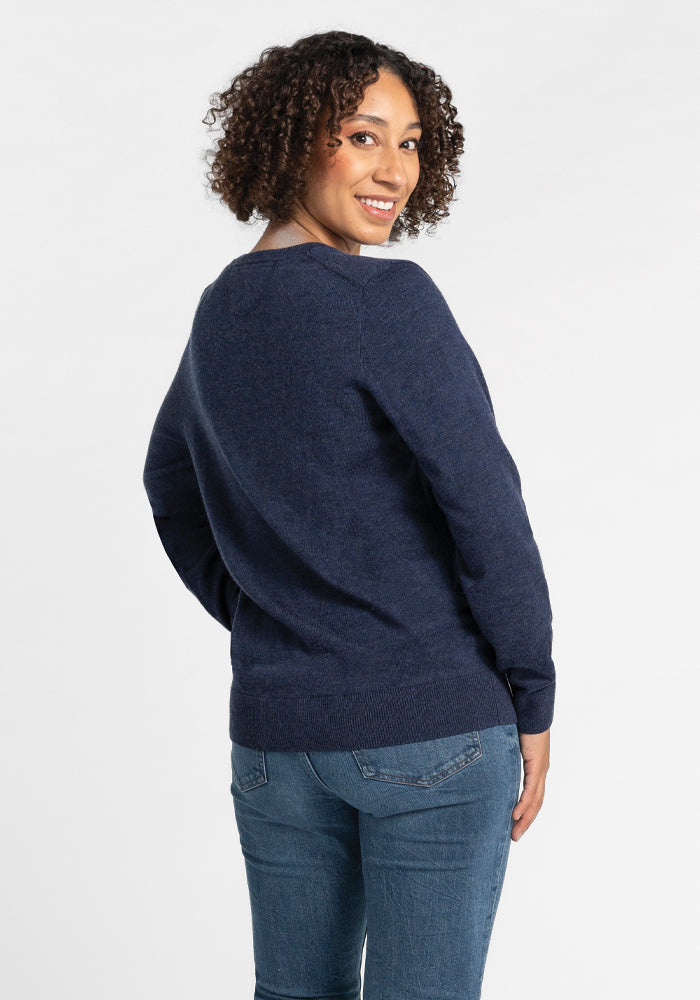 A person with curly hair is smiling and slightly turned to the side, wearing a Francesca Sweater in the Twilight Storm color by Woolx, paired with jeans, set against a plain light background.