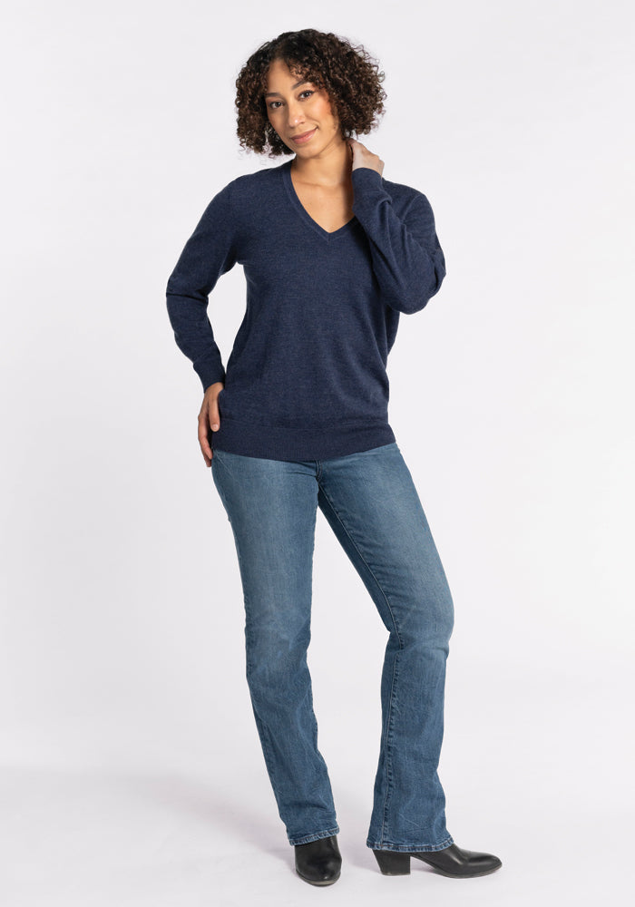 A person with curly hair is dressed in the luxurious Francesca Sweater in Twilight Storm by Woolx, along with blue jeans, posing with one hand on their hip and the other touching their face. They stand against a plain white background, wearing black shoes.