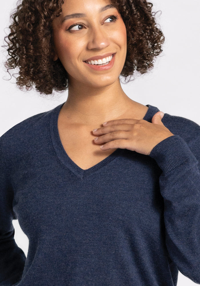 A person with curly hair beams with joy as they wear the Francesca Sweater in the elegant Twilight Storm color from Woolx. Made of luxurious Merino wool, this dark blue V-neck sweater highlights their hand resting on their chest. The simple white backdrop accentuates their happiness.