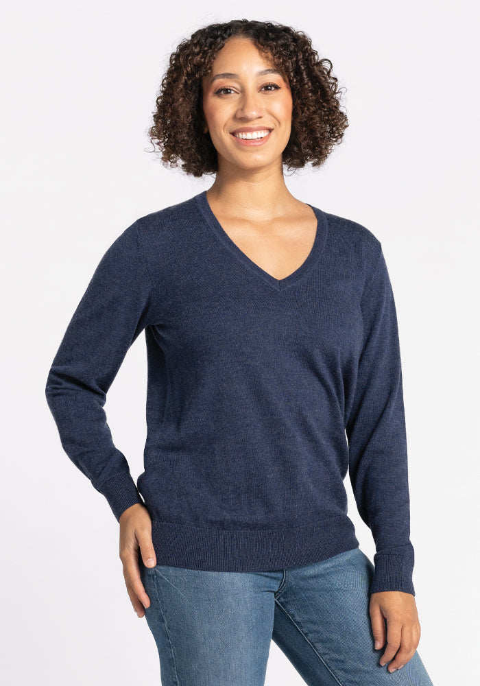 A person with curly hair is smiling and standing casually, wearing the Francesca Sweater in Twilight Storm by Woolx paired with blue jeans against a plain white background.