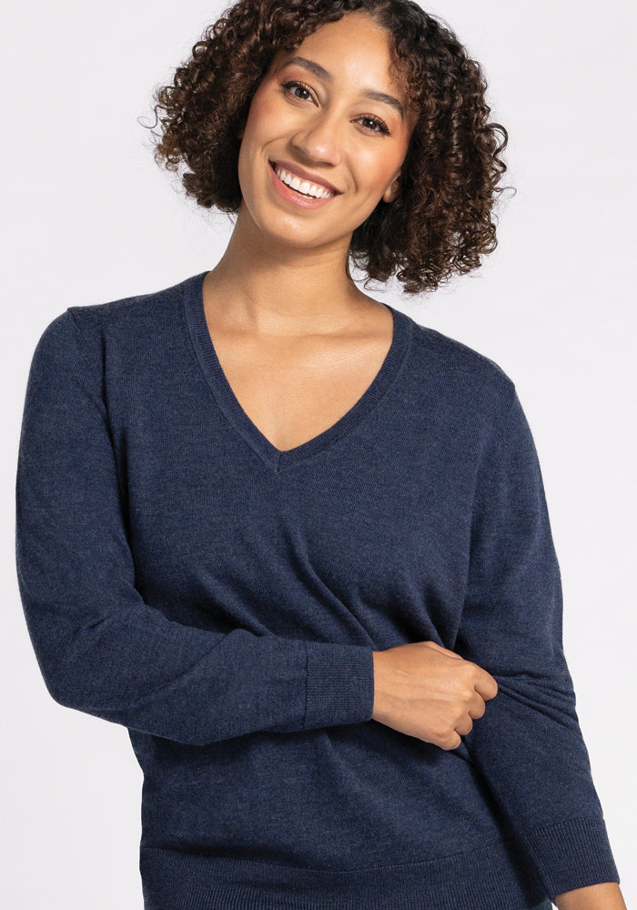 A person with curly hair smiles while wearing the elegant Francesca Sweater from Woolx in Twilight Storm, featuring a luxurious navy blue V-neck design crafted from the finest Merino wool. The background is plain and light-colored.