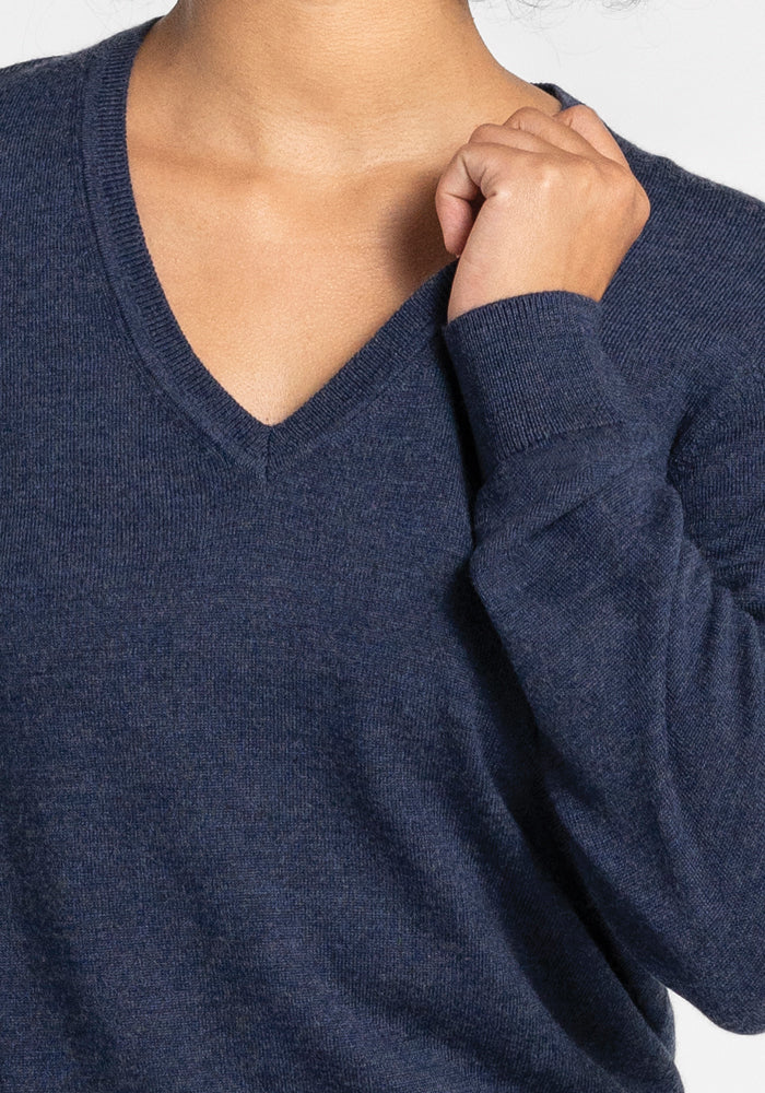 A person wearing the Woolx Francesca Sweater in the luxurious Twilight Storm shade gently holds the fabric near the collar. The image captures the upper torso, highlighting the texture of the Merino wool knit against a light gray background.