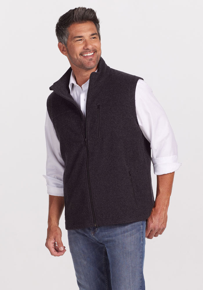 A man with short, dark hair is smiling and looking to the side. He is wearing a white button-up shirt with rolled-up sleeves and a warm Stowe Vest in Carbon Black by Woolx. He has on light blue jeans and is posed against a plain white background. 