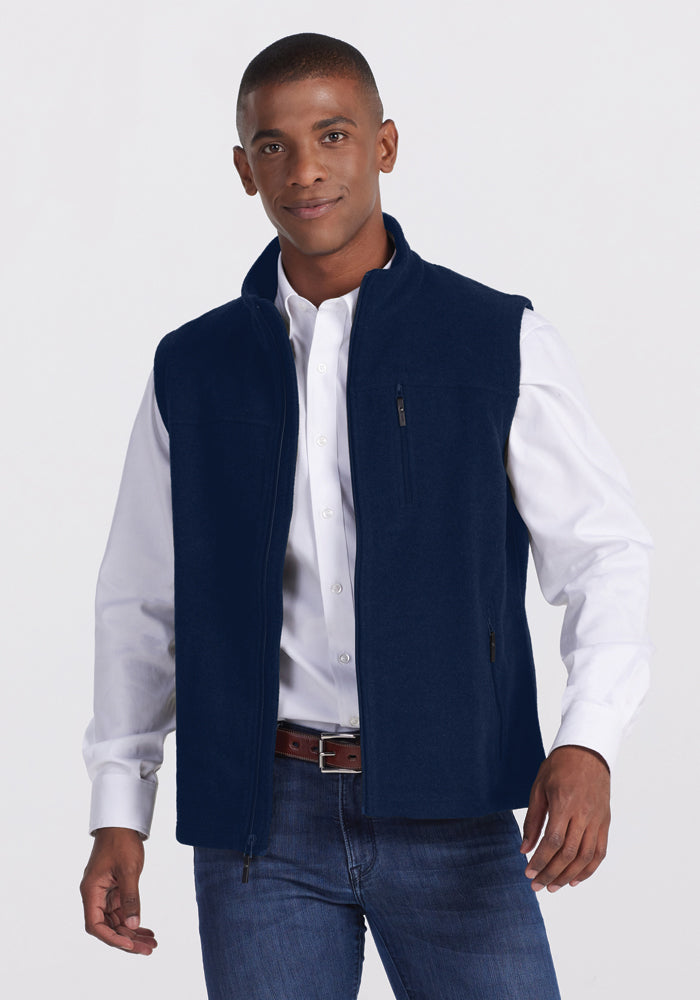 A man is standing and smiling, showcasing the Woolx Stowe Vest in navy blue over a white button-down shirt. He pairs it with blue jeans and a brown belt. The ultra-heavyweight Merino wool Stowe Vest serves as versatile outdoor gear, ideal for cold weather layering. The background is plain and light-colored. 