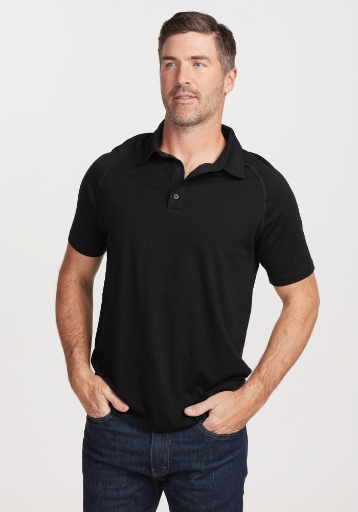 A man wearing the Summit Polo - Black by Woolx, renowned for its moisture management qualities, stands with his hands in his pockets. He has short hair and gazes slightly to the side. Paired with dark jeans, he exudes a relaxed demeanor against a simple, light-colored backdrop.