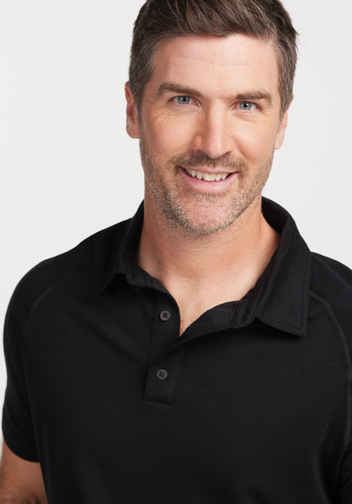 A smiling man with short hair and a light beard is wearing the Summit Polo - Black by Woolx, designed for moisture management. The background is plain white.