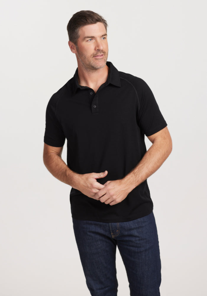 A man with short hair is wearing the Woolx Summit Polo in black, designed for moisture management, along with dark jeans. He looks to the side with a neutral expression against a plain white backdrop.