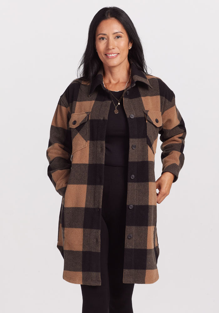 A person with long black hair is wearing the Sutton Shacket by Woolx, which features a camel checkered pattern, over a sleek black outfit. They are smiling with their hands in the shacket pockets while standing against a plain white background.