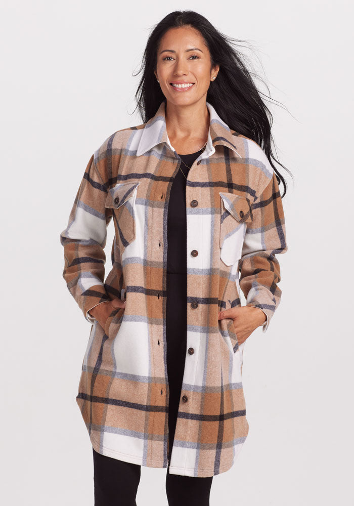 A woman is wearing a Woolx Sutton Shacket in Camel Plaid over a black shirt and black pants. She is smiling with her hands in the shacket's pockets. Perfect winter wear with its cozy look, her Sutton Shacket stands out against the plain white background.