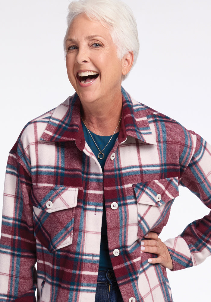 A smiling older woman with short white hair wears the Sutton Shacket in Frozen Cranberry Plaid from Woolx over a blue top—a perfect example of winter fashion. She has her right hand on her hip, showcasing a pendant necklace against the plain white background.