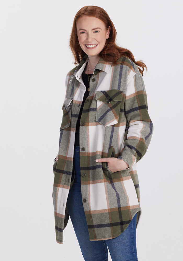 A person with long auburn hair is smiling and standing against a plain background. They are wearing the Sutton Shacket - Forest Plaid from Woolx, with their hands in the pockets, paired with blue jeans and a black top. 