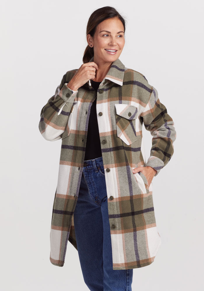 A woman wearing a long Woolx Sutton Shacket in Forest Plaid stylishly pairs it with a black top and blue jeans. Her left hand rests in her pocket while her right hand touches her chin as she smiles against a simple, light-colored background.