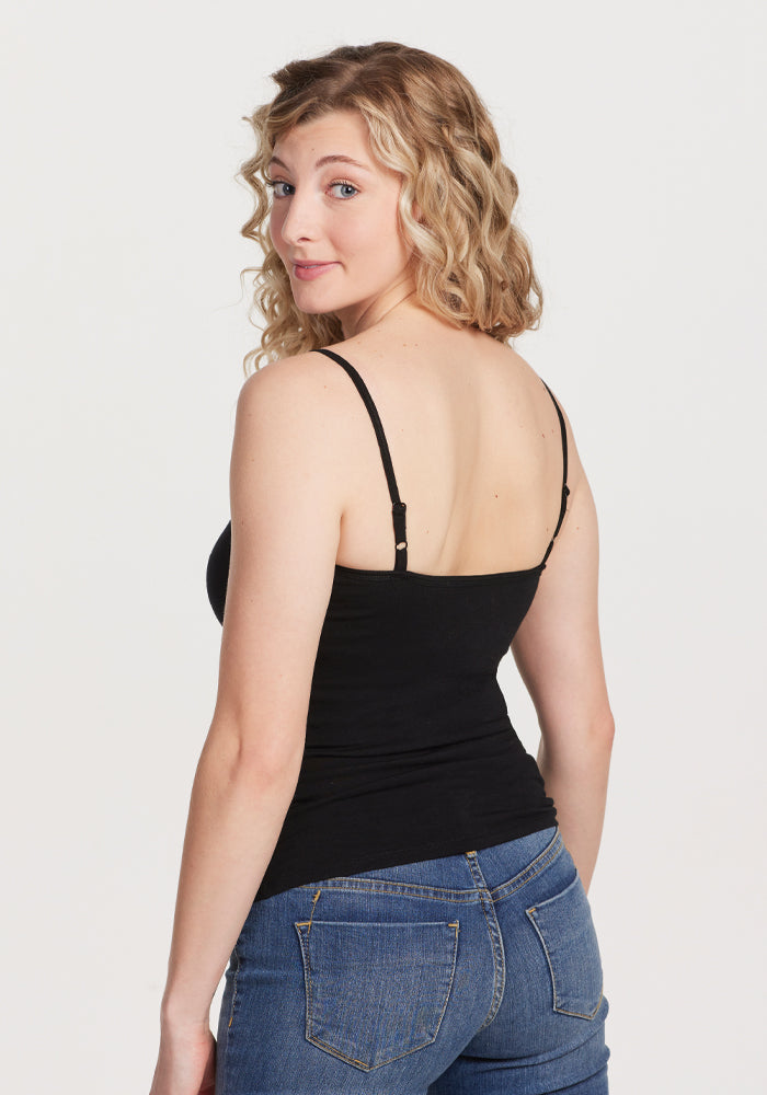 A woman with curly blonde hair faces backward, glancing over her shoulder. She wears the Woolx Teagen Cami With Shelf Bra - Black paired with blue jeans, creating a sleek and lightweight look against the plain white background.