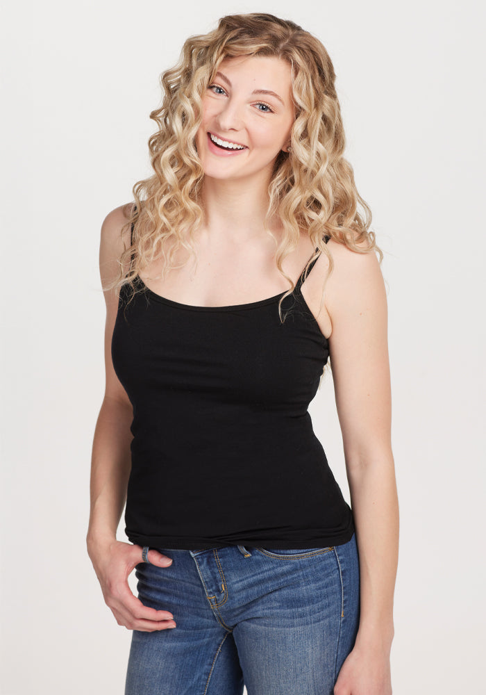 A smiling woman with long, curly blonde hair poses casually in a black Woolx Teagen cami featuring a built-in shelf bra and blue jeans against a plain white background, ideal for lightweight layering. 