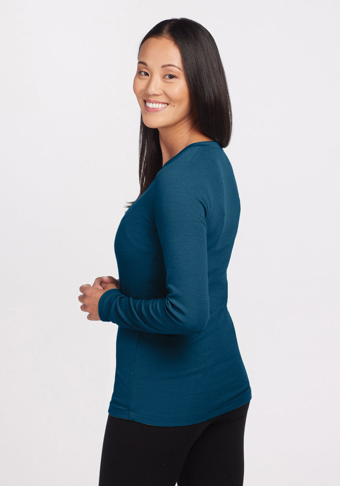 A person with long dark hair is smiling and posing in a side profile. They are wearing the Reese Ribbed Henley in Real Teal by Woolx and black pants, set against a plain white background.