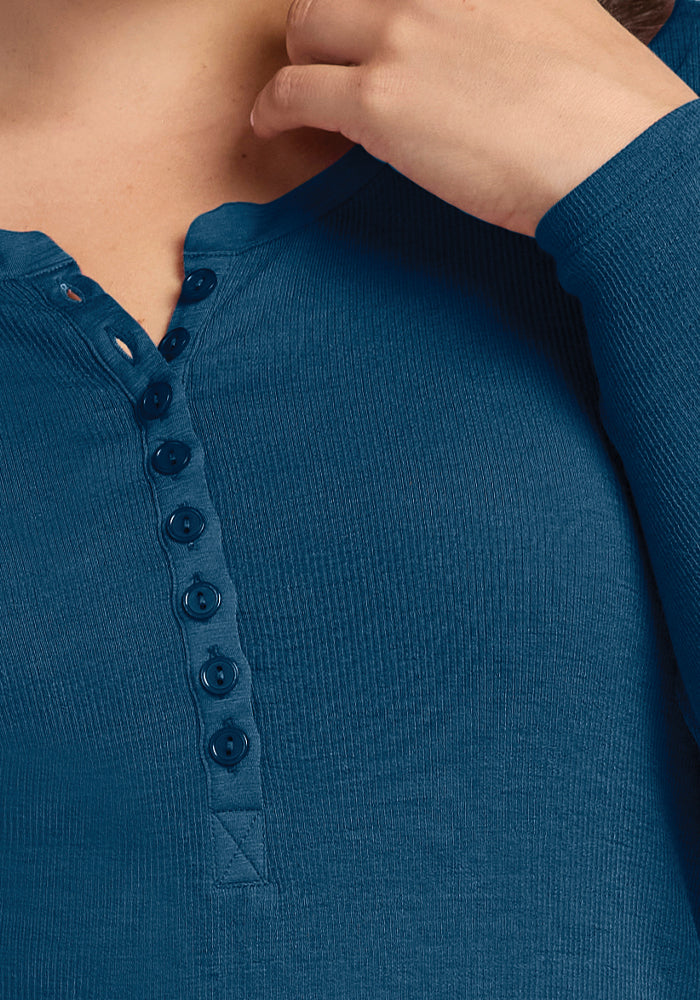 A person dressed in the Reese Ribbed Henley by Woolx in Real Teal, crafted from merino wool, with a series of buttons along the placket. They lift one hand to their neck, accentuating the texture and button details while enjoying its moisture-wicking comfort.