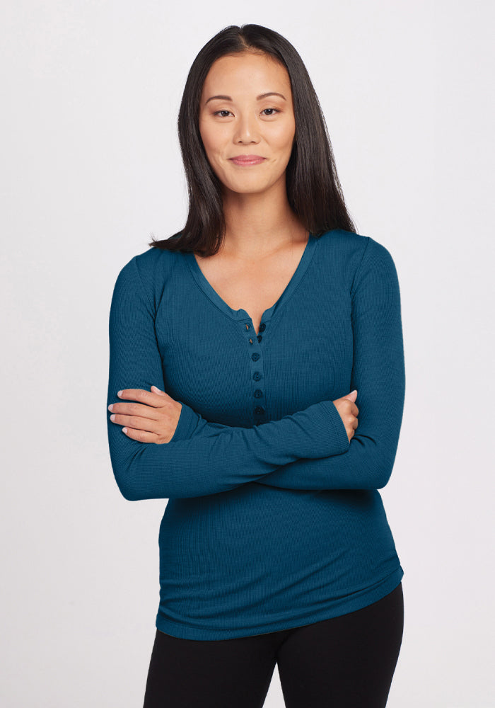 A person with long dark hair stands against a plain background, wearing the Reese Ribbed Henley in Real Teal from Woolx. They are smiling with arms crossed. 