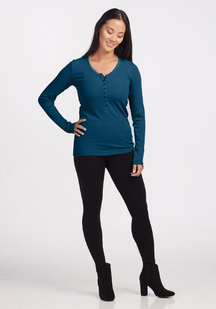 A person stands smiling, wearing the Reese Ribbed Henley in Real Teal from Woolx, paired with black leggings and black ankle boots. The Merino wool fabric ensures their comfort through its moisture-wicking properties. They strike a casual pose with one hand on the hip and the other by their side against a plain white background.