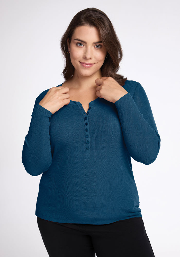 A person with long brown hair is wearing the Reese Ribbed Henley in Real Teal from Woolx, featuring buttons down the front and made of merino wool. They have paired it with black pants and are standing against a plain white background, holding the shirt collar with both hands while smiling gently at the camera. 