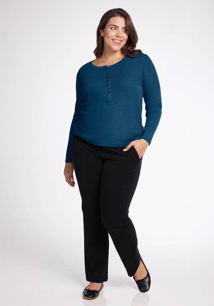 A woman wearing the Reese Ribbed Henley in Real Teal by Woolx and black pants stands smiling with one hand in her pocket. She is also wearing black shoes and is set against a plain white background.