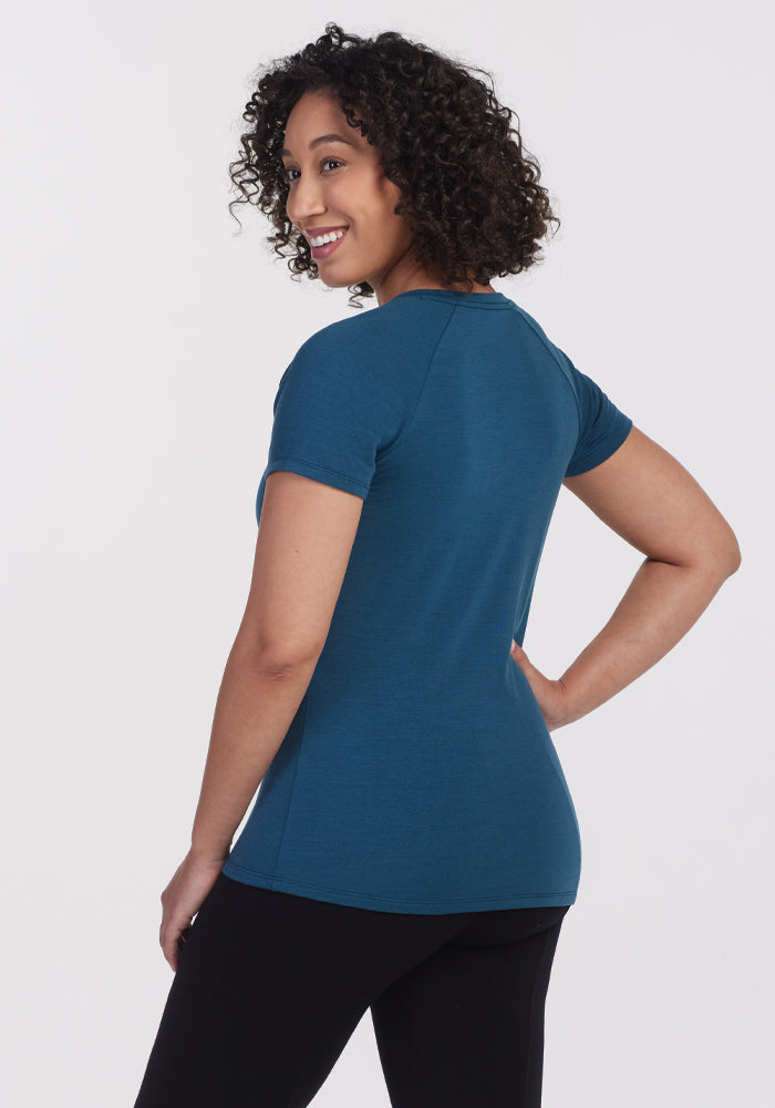 A person with curly hair is smiling and standing with one hand on their hip, wearing the Mia V Neck in Real Teal by Woolx and black pants, viewed from the side against a plain background.