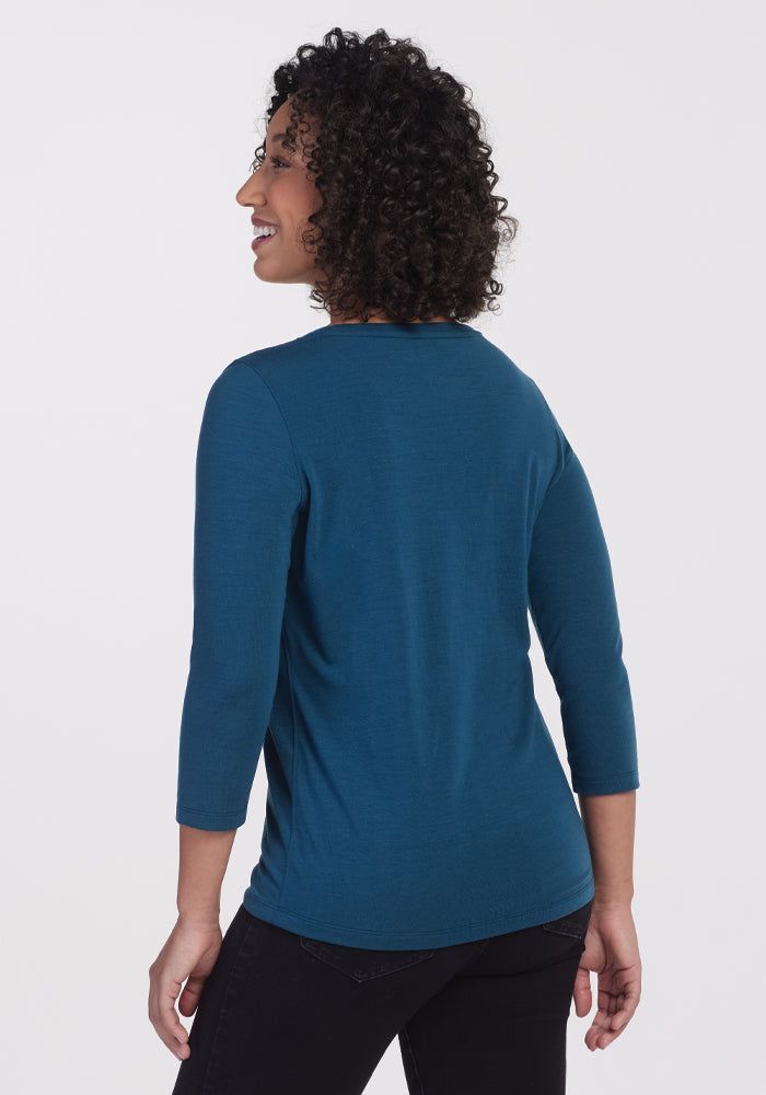 A person with curly hair is smiling and looking to the side. They are wearing the Jenny 3/4 Sleeve Crew Neck in Real Teal from Woolx, along with black pants made from DuraLite fabric. The background is plain white.