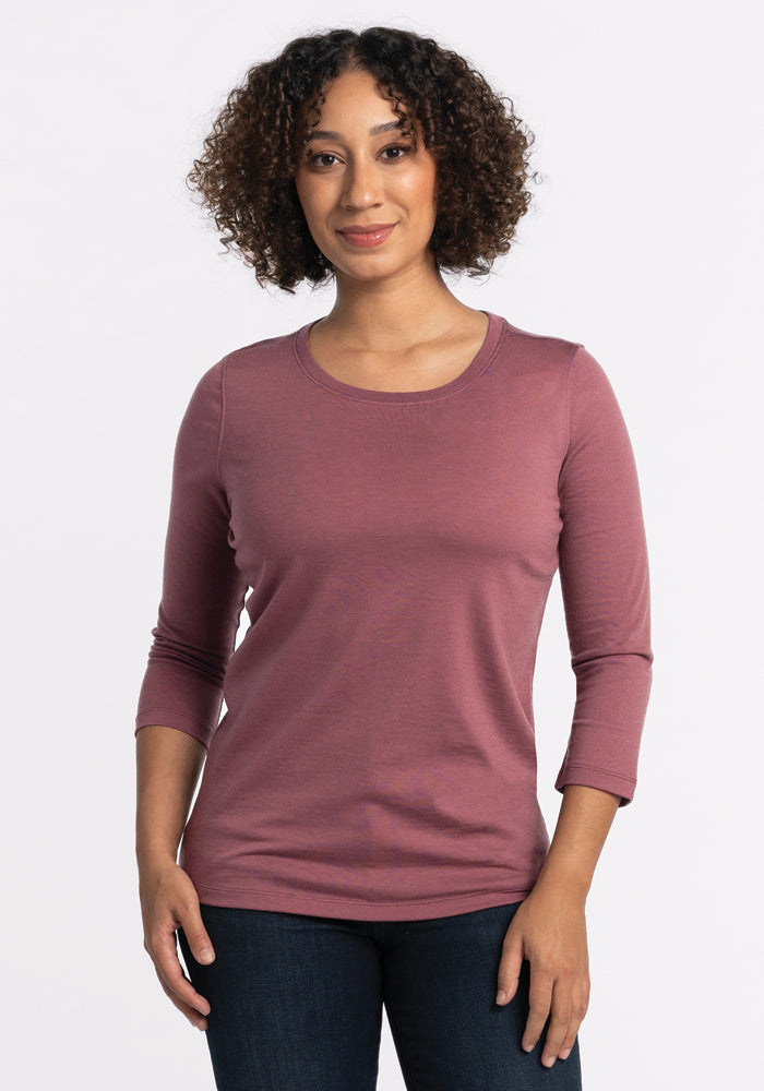 A person with curly hair is sporting a 3/4 sleeve, Wild Ginger Jenny Crew Neck by Woolx alongside dark jeans. They are standing against a plain white background and smiling gently. 