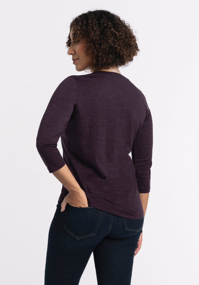 A person with curly hair is standing with their back to the camera, wearing a relaxed fit Elena V Neck - Deep Plum top by Woolx and dark jeans. The Merino wool top features three-quarter sleeves, and they have one hand on their hip against a plain white background.