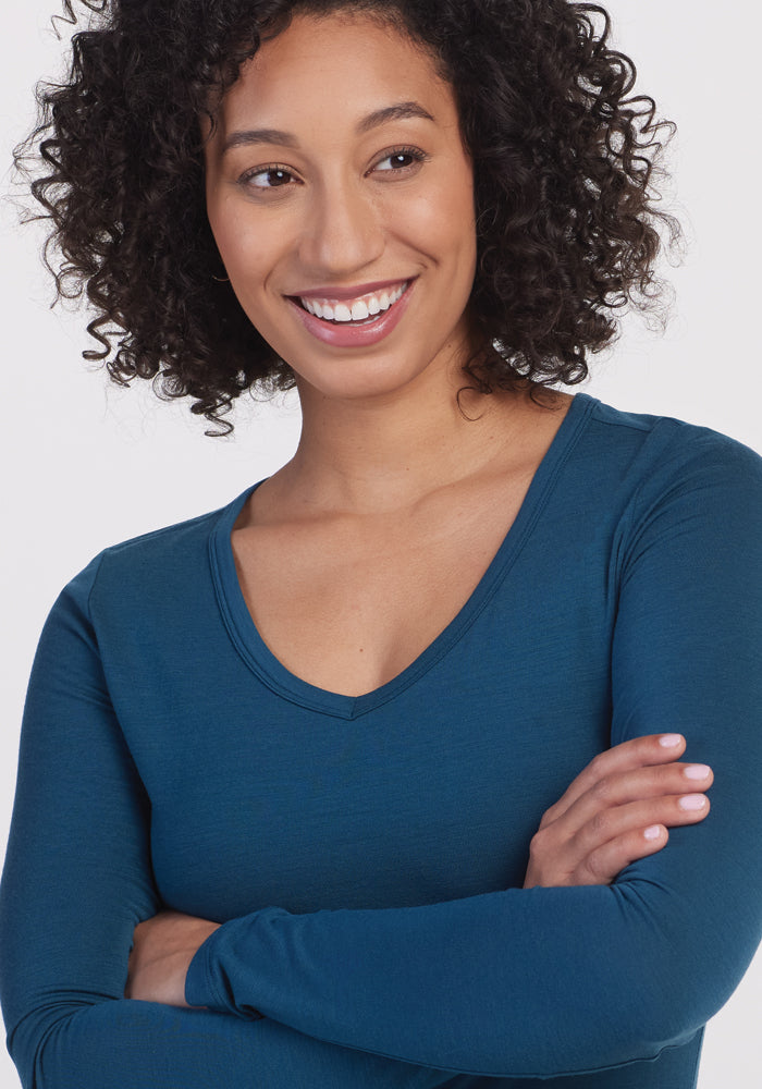 A person with curly hair smiles while looking to their side, wearing the Layla V Neck Top in Real Teal from Woolx, and has their arms crossed.