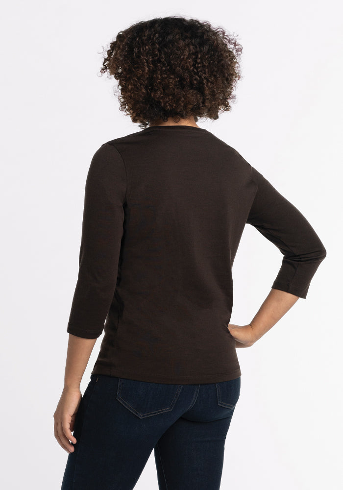 A person with curly hair is wearing a French Roast Elena V Neck from Woolx and dark jeans. They are facing away from the camera, exuding a modern chic vibe with their left hand on their hip against a plain white background.