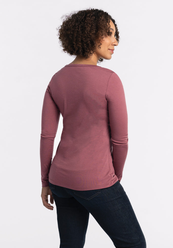 A person with curly hair is standing and looking to the side, wearing a Woolx Layla V Neck Top in wild ginger and dark jeans against a plain white background.