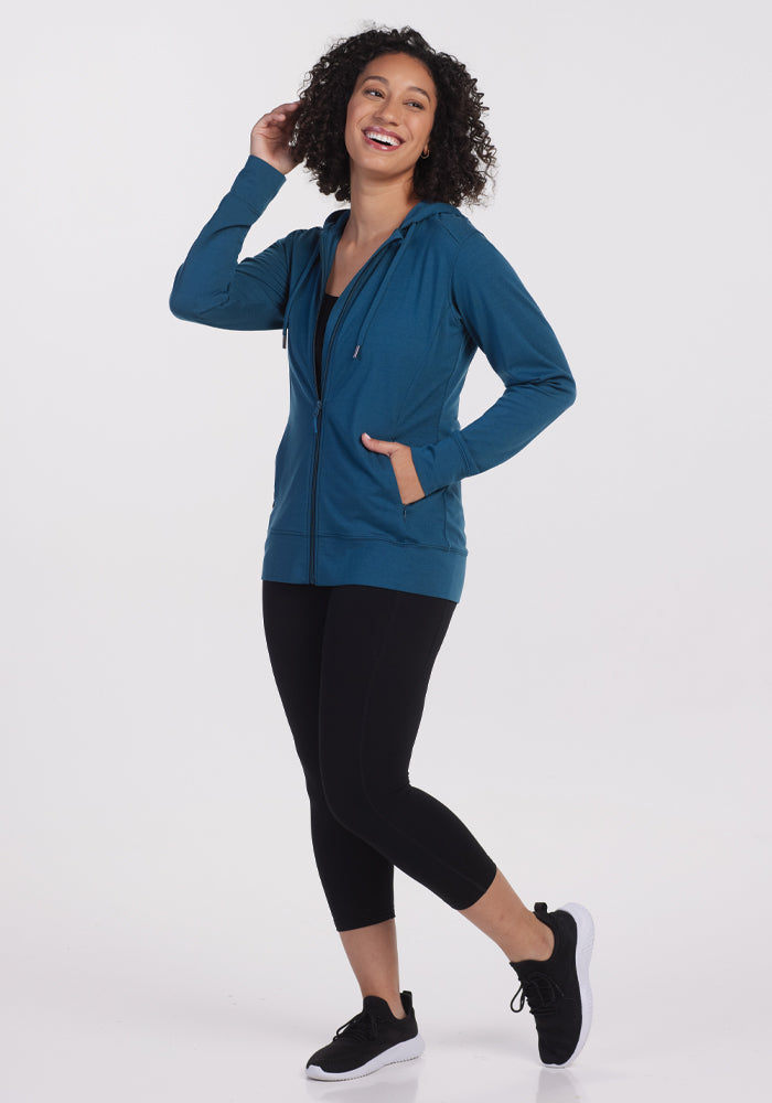 A person wearing the Ryann Hoodie in Real Teal from Woolx, paired with black capri leggings and black sneakers, is smiling and balancing on one leg against a plain white background. Their left hand is raised near their head, and the hoodie features pockets.