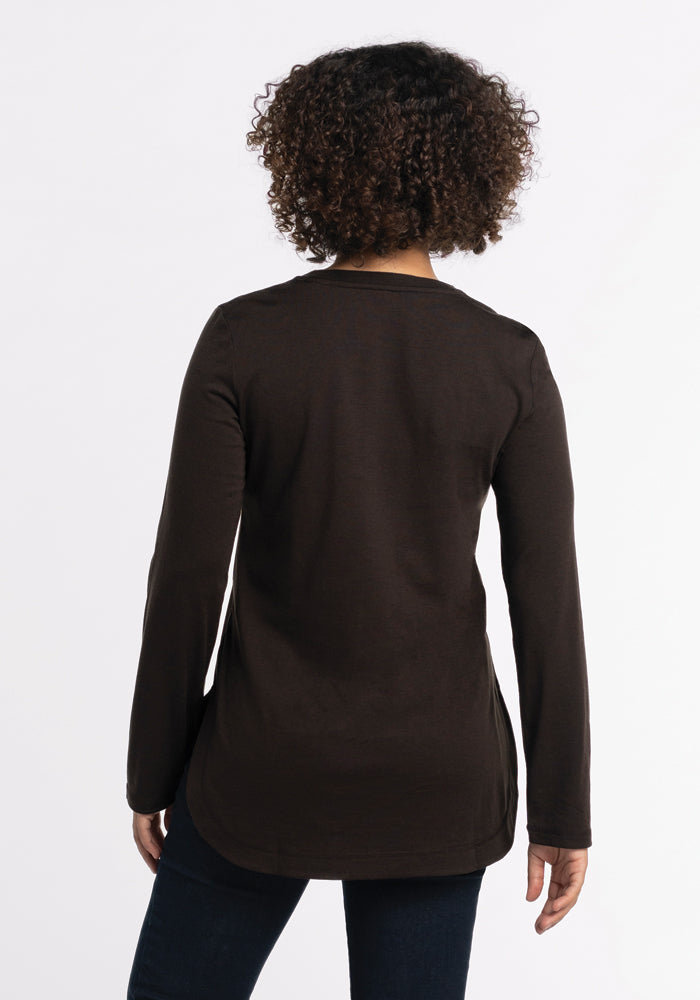 A person with curly hair is wearing a long-sleeved Woolx Hazel Tunic in French Roast, paired with dark pants, standing with their back to the camera against a plain white background.